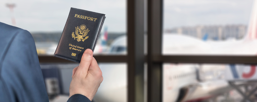 An immigrant at an airport holding up an American passport while looking out at an airplane.