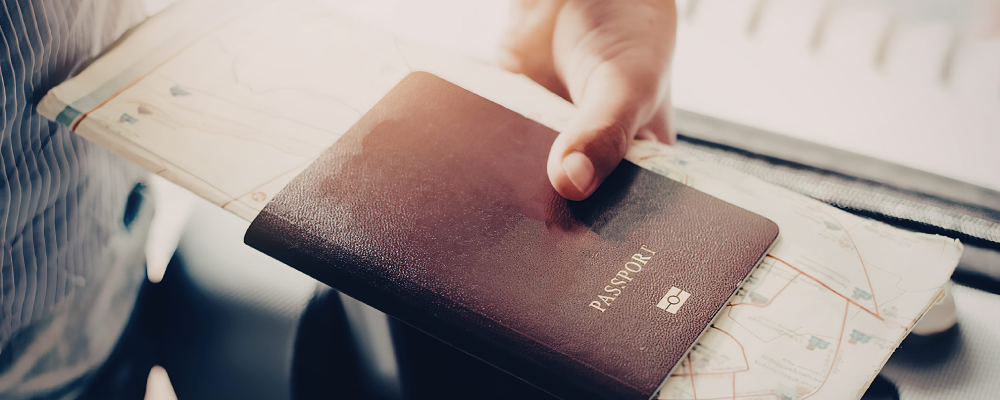 An immigrant's hand holding a passport and map while pulling a suitcase behind.